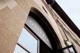 An upward view of a stone building facade featuring a large arched window. Above the window, there is signage displaying the words 'Gucci Osteria da Massimo Bottura'. The architectural design suggests a classic or traditional style with neutral tones.