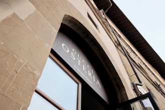 An upward view of a stone building facade featuring a large arched window. Above the window, there is signage displaying the words 'Gucci Osteria da Massimo Bottura'. The architectural design suggests a classic or traditional style with neutral tones.