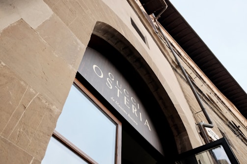 An upward view of a stone building facade featuring a large arched window. Above the window, there is signage displaying the words 'Gucci Osteria da Massimo Bottura'. The architectural design suggests a classic or traditional style with neutral tones.