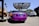 A vividly colored purple luxury convertible is parked at an auto center with a prominent sign above that reads 'RDB Five Star Tires Complete Auto Center'. Nearby, there are other vehicles including a white SUV and a blue sedan. An orange city bus is visible in the background on the left side next to some palm trees and graffiti art.