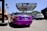 A vividly colored purple luxury convertible is parked at an auto center with a prominent sign above that reads 'RDB Five Star Tires Complete Auto Center'. Nearby, there are other vehicles including a white SUV and a blue sedan. An orange city bus is visible in the background on the left side next to some palm trees and graffiti art.