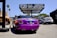 A vividly colored purple luxury convertible is parked at an auto center with a prominent sign above that reads 'RDB Five Star Tires Complete Auto Center'. Nearby, there are other vehicles including a white SUV and a blue sedan. An orange city bus is visible in the background on the left side next to some palm trees and graffiti art.