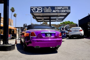 A vividly colored purple luxury convertible is parked at an auto center with a prominent sign above that reads 'RDB Five Star Tires Complete Auto Center'. Nearby, there are other vehicles including a white SUV and a blue sedan. An orange city bus is visible in the background on the left side next to some palm trees and graffiti art.
