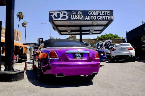 A vividly colored purple luxury convertible is parked at an auto center with a prominent sign above that reads 'RDB Five Star Tires Complete Auto Center'. Nearby, there are other vehicles including a white SUV and a blue sedan. An orange city bus is visible in the background on the left side next to some palm trees and graffiti art.