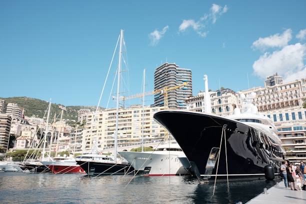 A luxurious marina lined with large yachts, nestled against a backdrop of modern and classic buildings. The sky is clear with a few clouds, and the foreground features a shiny black yacht prominently docked by a pier with people nearby.