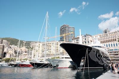 A luxurious marina lined with large yachts, nestled against a backdrop of modern and classic buildings. The sky is clear with a few clouds, and the foreground features a shiny black yacht prominently docked by a pier with people nearby.