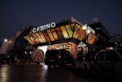 A building with a brightly illuminated entrance is seen at night. The sign above reads 'Casino' in bright letters. The facade features colorful graphics depicting various indulgent themes. Several vehicles are parked near the entrance, and people are visible in the vicinity. Tall lamp posts with spherical lights are present, casting a warm glow.