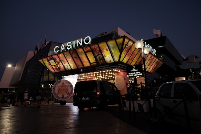 A building with a brightly illuminated entrance is seen at night. The sign above reads 'Casino' in bright letters. The facade features colorful graphics depicting various indulgent themes. Several vehicles are parked near the entrance, and people are visible in the vicinity. Tall lamp posts with spherical lights are present, casting a warm glow.