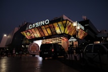 A building with a brightly illuminated entrance is seen at night. The sign above reads 'Casino' in bright letters. The facade features colorful graphics depicting various indulgent themes. Several vehicles are parked near the entrance, and people are visible in the vicinity. Tall lamp posts with spherical lights are present, casting a warm glow.