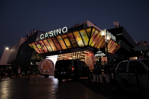 A building with a brightly illuminated entrance is seen at night. The sign above reads 'Casino' in bright letters. The facade features colorful graphics depicting various indulgent themes. Several vehicles are parked near the entrance, and people are visible in the vicinity. Tall lamp posts with spherical lights are present, casting a warm glow.