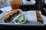 A tray holding two halves of a grilled sandwich, with one half showing pulled meat and greens spilling from crusty bread. Accompanying the sandwich is a small cup of pickled cucumber slices. In the background, an iced drink in a clear cup displays the text 'JOAN'S ON THIRD'. The setting appears to be a casual dining environment.