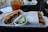 A tray holding two halves of a grilled sandwich, with one half showing pulled meat and greens spilling from crusty bread. Accompanying the sandwich is a small cup of pickled cucumber slices. In the background, an iced drink in a clear cup displays the text 'JOAN'S ON THIRD'. The setting appears to be a casual dining environment.