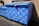 A large blue dumpster is decorated with pink graffiti patterns resembling interconnected loops. The design is repeated across the surface of the dumpster, which is situated outdoors near a building's wall. Sunlight casts shadows, highlighting the texture of the blue paint.