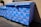 A large blue dumpster is decorated with pink graffiti patterns resembling interconnected loops. The design is repeated across the surface of the dumpster, which is situated outdoors near a building's wall. Sunlight casts shadows, highlighting the texture of the blue paint.