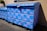 A large blue dumpster is decorated with pink graffiti patterns resembling interconnected loops. The design is repeated across the surface of the dumpster, which is situated outdoors near a building's wall. Sunlight casts shadows, highlighting the texture of the blue paint.