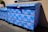 A large blue dumpster is decorated with pink graffiti patterns resembling interconnected loops. The design is repeated across the surface of the dumpster, which is situated outdoors near a building's wall. Sunlight casts shadows, highlighting the texture of the blue paint.