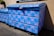 A large blue dumpster is decorated with pink graffiti patterns resembling interconnected loops. The design is repeated across the surface of the dumpster, which is situated outdoors near a building's wall. Sunlight casts shadows, highlighting the texture of the blue paint.