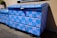 A large blue dumpster is decorated with pink graffiti patterns resembling interconnected loops. The design is repeated across the surface of the dumpster, which is situated outdoors near a building's wall. Sunlight casts shadows, highlighting the texture of the blue paint.