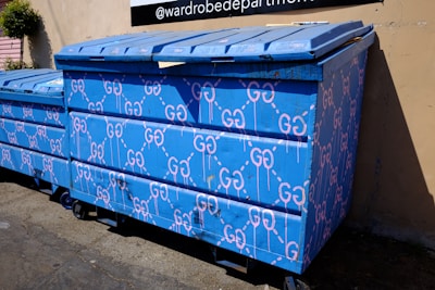 A large blue dumpster is decorated with pink graffiti patterns resembling interconnected loops. The design is repeated across the surface of the dumpster, which is situated outdoors near a building's wall. Sunlight casts shadows, highlighting the texture of the blue paint.