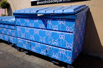 A large blue dumpster is decorated with pink graffiti patterns resembling interconnected loops. The design is repeated across the surface of the dumpster, which is situated outdoors near a building's wall. Sunlight casts shadows, highlighting the texture of the blue paint.