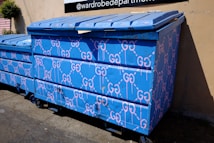 A large blue dumpster is decorated with pink graffiti patterns resembling interconnected loops. The design is repeated across the surface of the dumpster, which is situated outdoors near a building's wall. Sunlight casts shadows, highlighting the texture of the blue paint.