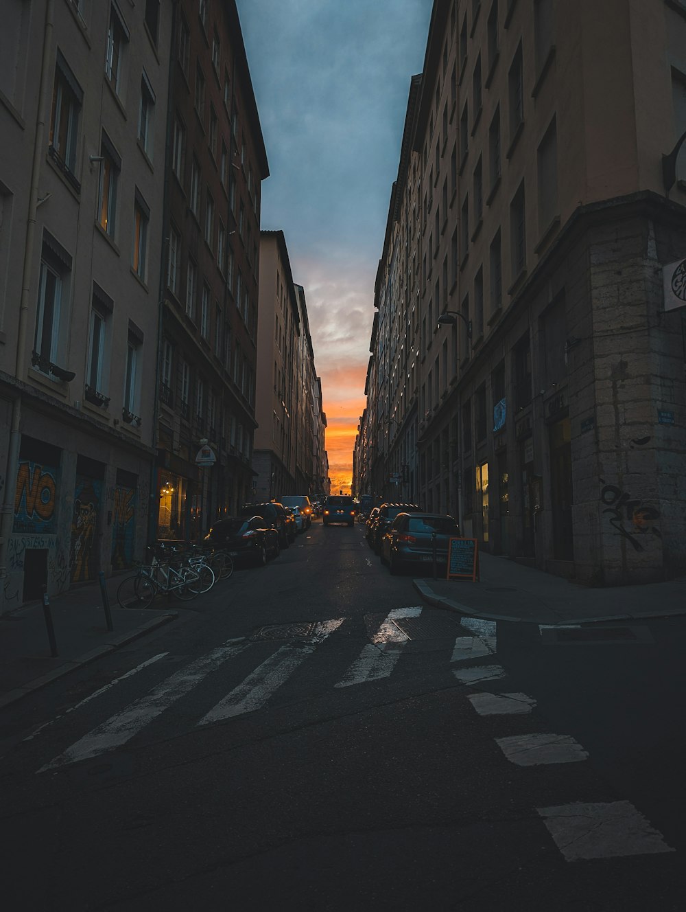 a city street with tall buildings and a sunset in the background