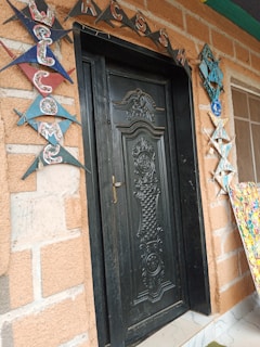 A black, ornately carved door set in a brick wall, decorated with colorful triangular flags spelling 'WELCOME' on the left side. Above the door, the letters 'ACCESS' are arranged in a decorative pattern. To the right, there is a decorative hanging and a partially visible colorful abstract art piece.