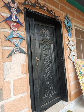 A black, ornately carved door set in a brick wall, decorated with colorful triangular flags spelling 'WELCOME' on the left side. Above the door, the letters 'ACCESS' are arranged in a decorative pattern. To the right, there is a decorative hanging and a partially visible colorful abstract art piece.