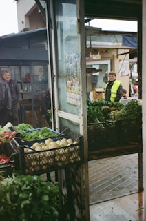 A bustling outdoor market setting with crates of fresh vegetables, including radishes and greens, displayed prominently. People are visible in the background, dressed warmly, suggesting a cool day. The structure is open, with visible signs and a calendar, evoking a casual and lively market atmosphere.
