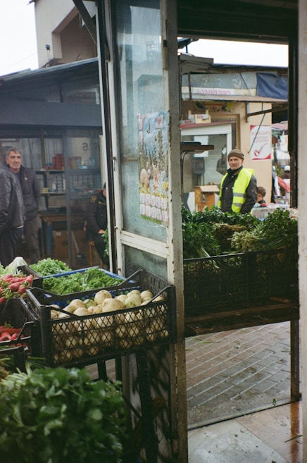 A bustling outdoor market setting with crates of fresh vegetables, including radishes and greens, displayed prominently. People are visible in the background, dressed warmly, suggesting a cool day. The structure is open, with visible signs and a calendar, evoking a casual and lively market atmosphere.