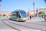 A modern tram moving along tracks in an urban area. The background features colorful buildings with large windows and people walking nearby. Streetlamps and traffic signs are visible, and the sky is clear.