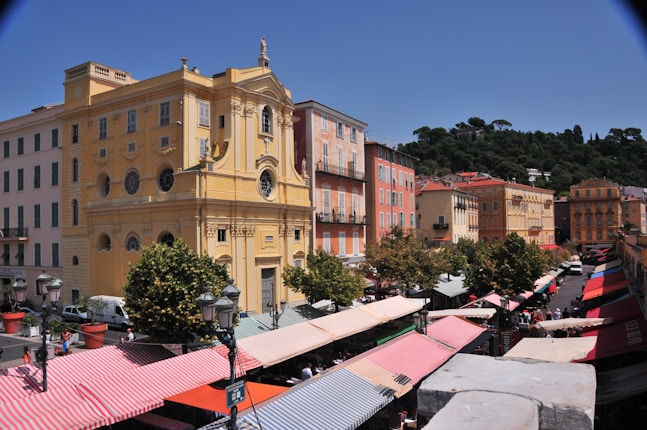 A vibrant street scene featuring colorful historic buildings lining a market square. The ornate yellow building stands out with architectural detailing, including circular windows and decorative elements. Market stalls with striped awnings add bursts of red, white, and pastel hues, while mature trees provide shaded areas. The lush green hillside in the background contrasts with the bright blue sky, enhancing the lively atmosphere.