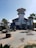 A coastal building with a circular tower and several palm trees nearby. The structure features a staircase leading up to a balcony, and artistic metal sculptures are placed in front of it. The building has a Mediterranean architectural style and the sky is clear.
