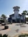 A coastal building with a circular tower and several palm trees nearby. The structure features a staircase leading up to a balcony, and artistic metal sculptures are placed in front of it. The building has a Mediterranean architectural style and the sky is clear.