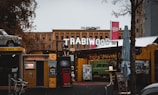 An outdoor urban scene featuring a kiosk with a 'Photoautomat' sign beside digital screens. A sign above reads 'TRABIWOOD' with a vintage car model placed on a roof. The area has a rustic, retro vibe with signs for a cinema and Berlin helicopter tours. Various trees and buildings frame the background, suggesting a lively urban environment.