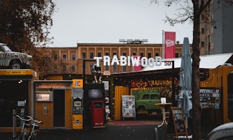 An outdoor urban scene featuring a kiosk with a 'Photoautomat' sign beside digital screens. A sign above reads 'TRABIWOOD' with a vintage car model placed on a roof. The area has a rustic, retro vibe with signs for a cinema and Berlin helicopter tours. Various trees and buildings frame the background, suggesting a lively urban environment.