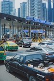 A busy toll booth area with multiple lanes, cars lined up, and overhead blue signs labeled 'Easy Pass' in Thai and English. Skyscrapers are visible in the background, indicating an urban setting. Vehicles of various types, including a black pickup truck, a green and yellow taxi, and several sedans, are present.
