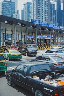 A busy toll booth area with multiple lanes, cars lined up, and overhead blue signs labeled 'Easy Pass' in Thai and English. Skyscrapers are visible in the background, indicating an urban setting. Vehicles of various types, including a black pickup truck, a green and yellow taxi, and several sedans, are present.