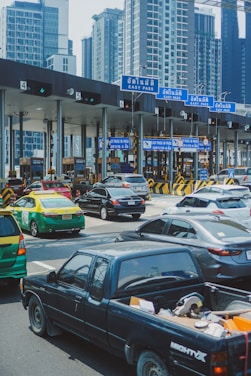 A busy toll booth area with multiple lanes, cars lined up, and overhead blue signs labeled 'Easy Pass' in Thai and English. Skyscrapers are visible in the background, indicating an urban setting. Vehicles of various types, including a black pickup truck, a green and yellow taxi, and several sedans, are present.