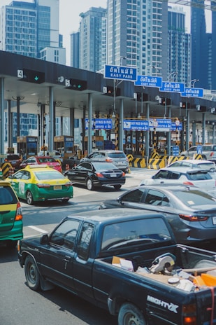 A busy toll booth area with multiple lanes, cars lined up, and overhead blue signs labeled 'Easy Pass' in Thai and English. Skyscrapers are visible in the background, indicating an urban setting. Vehicles of various types, including a black pickup truck, a green and yellow taxi, and several sedans, are present.