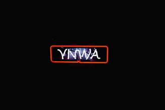 A neon sign with the letters 'YNWA' illuminated in white against a dark background. The sign is encased in a red rectangular outline, which provides contrast to the darkness surrounding it, making the text stand out distinctly.