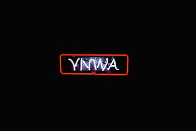 A neon sign with the letters 'YNWA' illuminated in white against a dark background. The sign is encased in a red rectangular outline, which provides contrast to the darkness surrounding it, making the text stand out distinctly.
