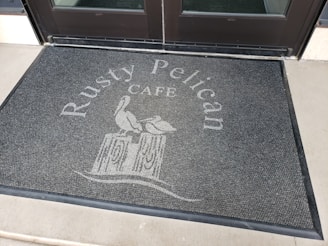A doormat placed in front of a glass door features the text 'Rusty Pelican Cafe' with an illustration of a pelican perched on a post.