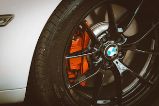 A close-up view of a black alloy wheel on a white car, featuring a visible silver brake disc and an orange brake caliper. The glossy finish of the wheel gives an impression of high quality and performance.