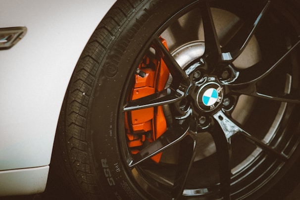 A close-up view of a black alloy wheel on a white car, featuring a visible silver brake disc and an orange brake caliper. The glossy finish of the wheel gives an impression of high quality and performance.