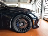 A close-up view of a sleek, black sports car with a focus on the front wheel and tire. The alloy rims are intricate and high-performance, featuring branding in the center. The car is parked on a polished, reddish-brown tile floor inside a modern garage with large windows.