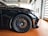 A close-up view of a sleek, black sports car with a focus on the front wheel and tire. The alloy rims are intricate and high-performance, featuring branding in the center. The car is parked on a polished, reddish-brown tile floor inside a modern garage with large windows.