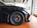 A close-up view of a sleek, black sports car with a focus on the front wheel and tire. The alloy rims are intricate and high-performance, featuring branding in the center. The car is parked on a polished, reddish-brown tile floor inside a modern garage with large windows.