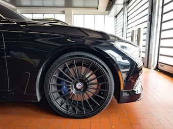 A close-up view of a sleek, black sports car with a focus on the front wheel and tire. The alloy rims are intricate and high-performance, featuring branding in the center. The car is parked on a polished, reddish-brown tile floor inside a modern garage with large windows.