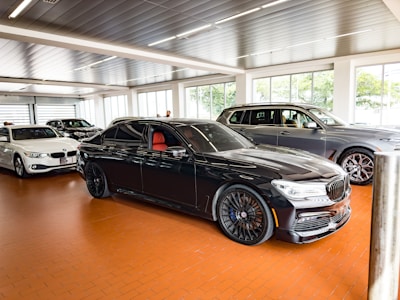 A showroom displaying several luxury cars, with a prominent black sedan featuring red interior seating in the foreground. The car is positioned on a polished brown tile floor under bright overhead lighting. Other vehicles, including a white sedan and a gray SUV, are aligned in the background in front of large windows that allow natural light to flood the room.
